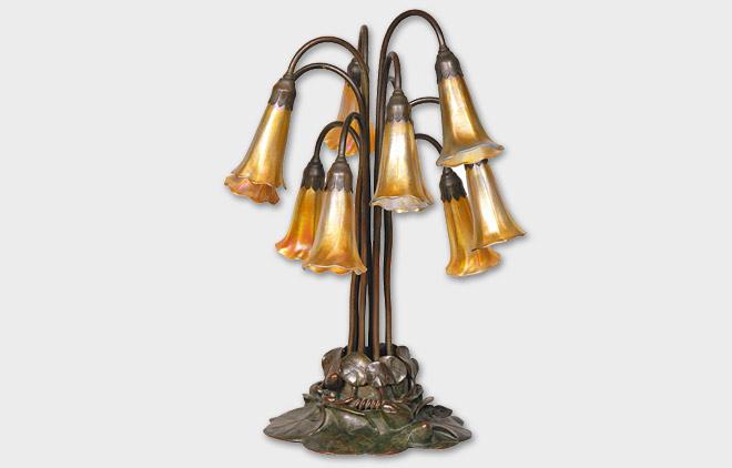 Louis Comfort Tiffany Bedeutende Tischlampe "Lily Pond" New York/USA, um 1900. Tischlampe mit 10 irisierenden Glasschirmen aus sog. Favrile-Glas, ätzsign. "LCT" und "Favrile". Bronzefuß, grünlich braun patiniert, Bodenplatte gest. und num. "28626 Tiffany Studios New York 3" und ligierts Monogr. "TGDCo". Der Bronzefuß in Form von Seerosenblättern mit 10 aufsteigenden Stengeln, die die goldgelb irisierenden Lampenschirme in Form von Lilienblüten tragen. Zuschlag: 28.000 €